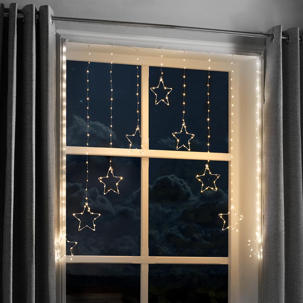 Warm white LED curtain string lights with hanging star-shaped bulbs draped over a window at night, framed by gray curtains.