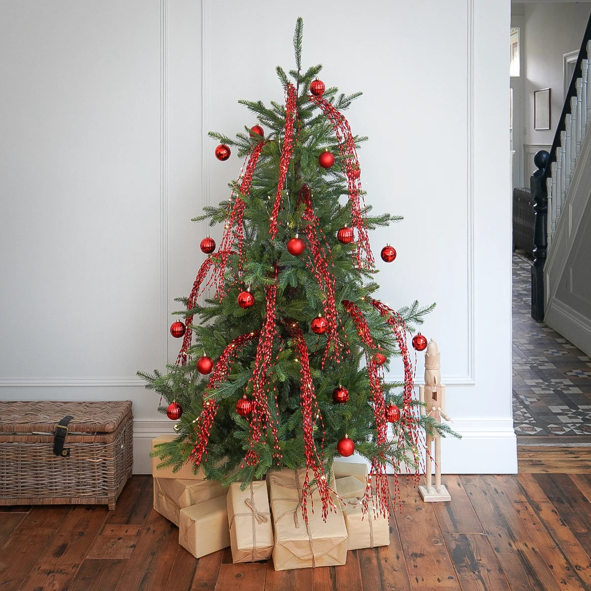 A decorated Christmas tree stands on wrapped gifts in a home interior with hardwood floors and a staircase visible in the background. The tree is green with red ornaments, red garlands, and warm white string lights.