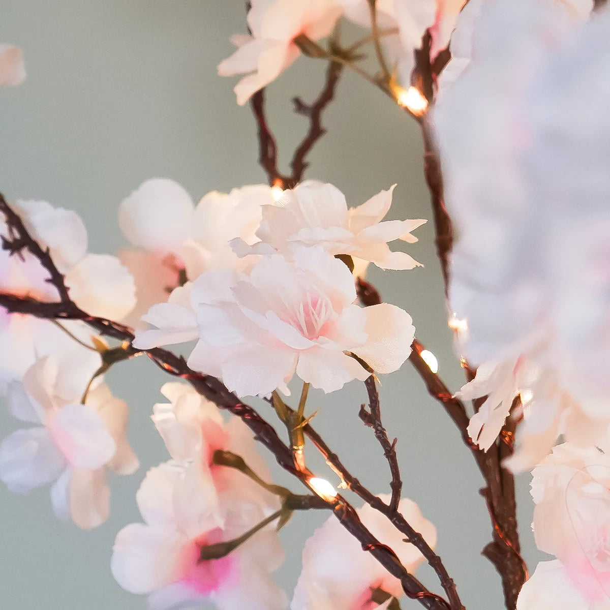 Lighted artificial cherry blossom branches with soft pink and white flowers on dark brown stems, decorated with small warm white string lights against a soft gray background.