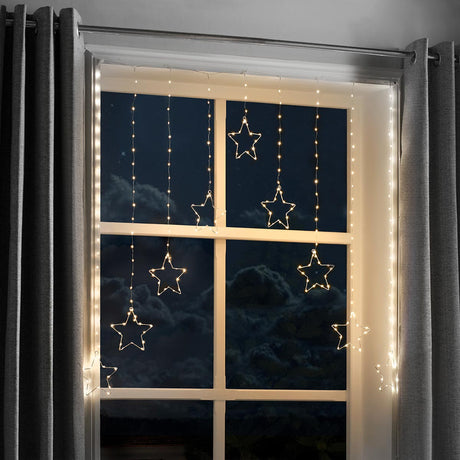 Warm white LED curtain string lights with hanging star-shaped bulbs draped over a window at night, framed by gray curtains.