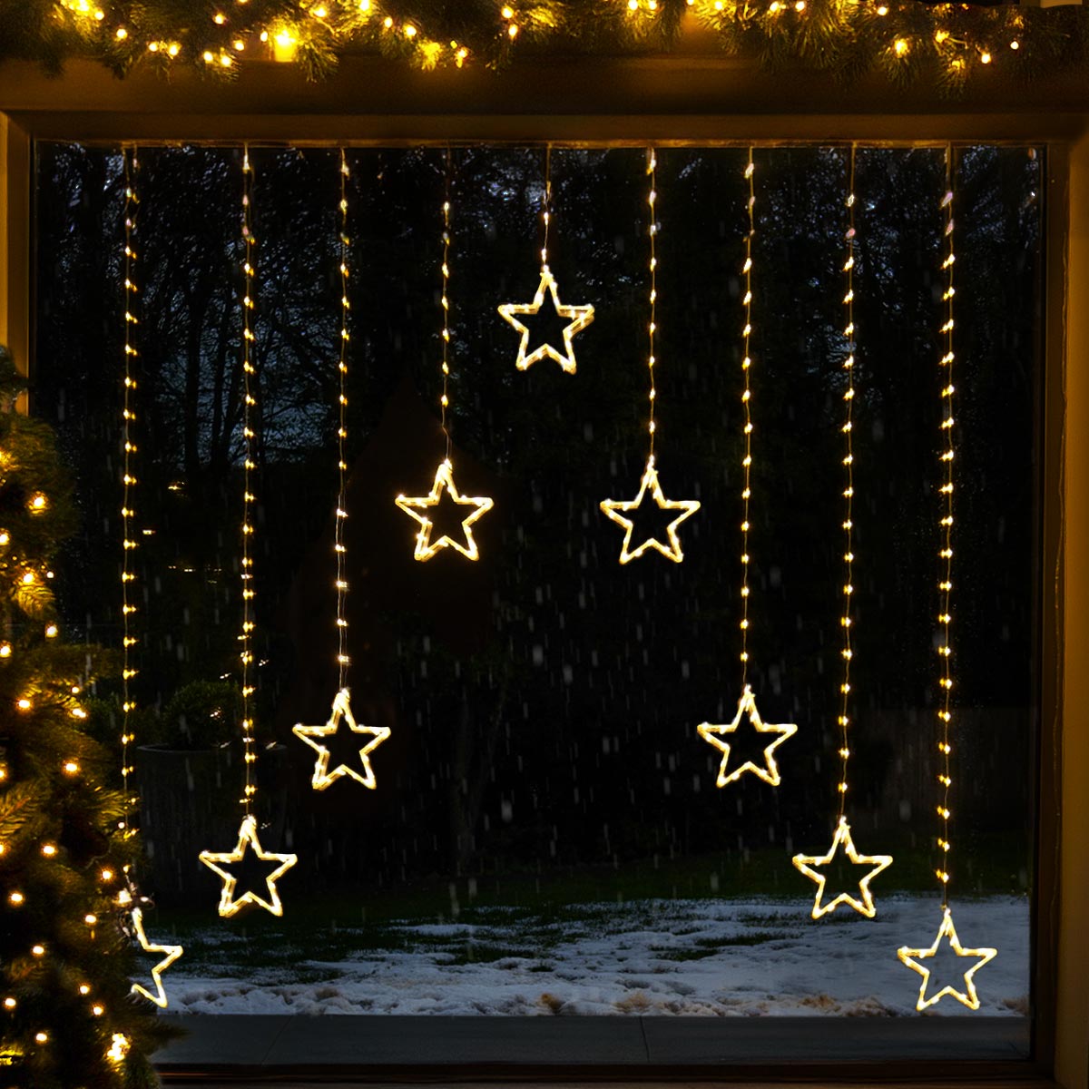 Warm white LED curtain string lights shaped like stars, hanging vertically in multiple strands across a christmas window during a snowy night scene.