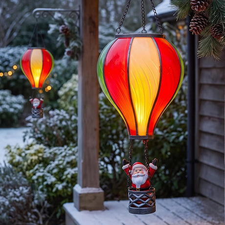 A hanging Christmas ornament shaped like a hot air balloon with a glowing red, orange, and yellow glass shade and a small Santa figure in a basket below, shown outdoors on a porch with a second matching ornament in the background.