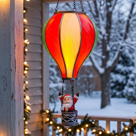 A hanging stained-glass ornament shaped like a hot air balloon with a Santa Claus figure in the basket, shown outdoors on a porch at night with warm string lights and a snowy yard in the background.