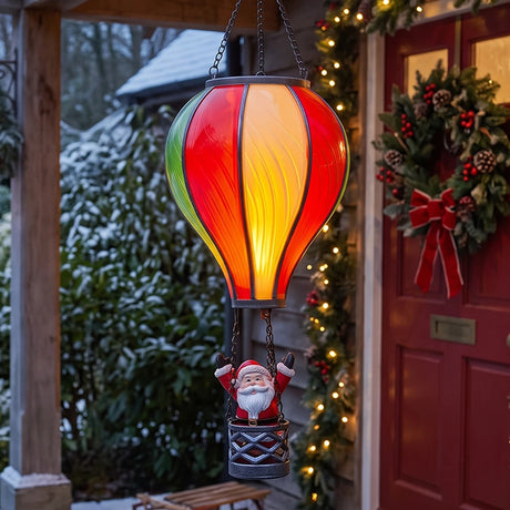 A hanging decorative lantern shaped like a hot air balloon with a Santa Claus figure in the basket, displayed in front of a decorated red holiday door.