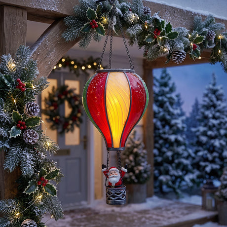 A festive Christmas hanging ornament shaped like a hot air balloon with a glowing yellow and red interior and a small Santa Claus figure in the basket, displayed outdoors under a decorated wooden porch arch with snowy trees in the background.