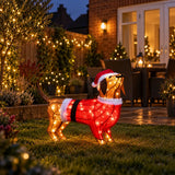 A wire-frame dachshund Christmas decoration wearing a Santa hat and red sweater with warm white lights, displayed on a lawn in a decorated backyard at night.