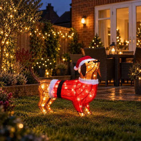 A wire-frame dachshund Christmas decoration wearing a Santa hat and red sweater with warm white lights, displayed on a lawn in a decorated backyard at night.