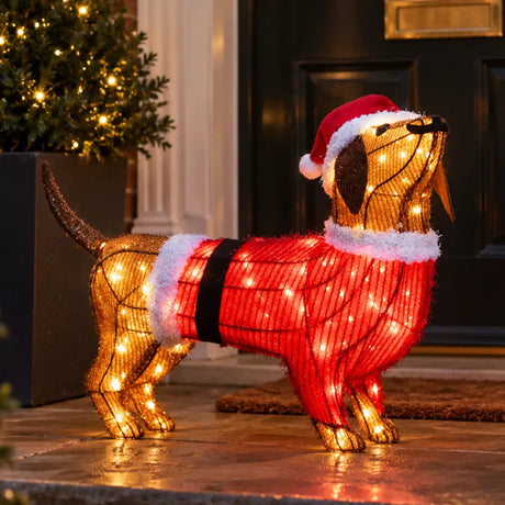 A wire-frame dachshund holiday decoration wearing a red Santa sweater with white faux-fur trim and a red Santa hat, lit with warm white lights, displayed on a front porch.