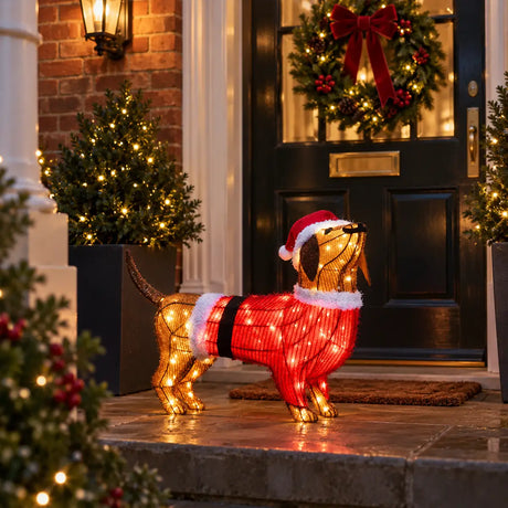 A festive dachshund-shaped light decoration wearing a red Santa hat and red sweater with white trim, displayed on a front porch in front of a decorated dark door with a wreath.