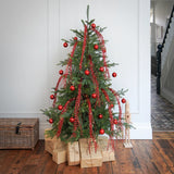 A decorated Christmas tree stands on wrapped gifts in a home interior with hardwood floors and a staircase visible in the background. The tree is green with red ornaments, red garlands, and warm white string lights.