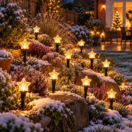 Outdoor star-shaped solar string lights glowing in a snowy garden landscape at night, mounted on rocks and plants with a decorated Christmas tree visible in the background.