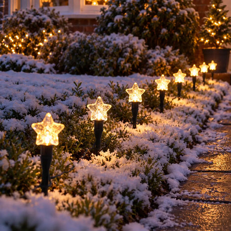 Outdoor star-shaped solar landscape lights glowing on snowy garden beds beside a walkway at night.