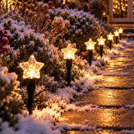 A row of glowing star-shaped outdoor lights mounted on black posts along a snowy garden path at dusk, with snow-covered shrubs and warm lights in the background.