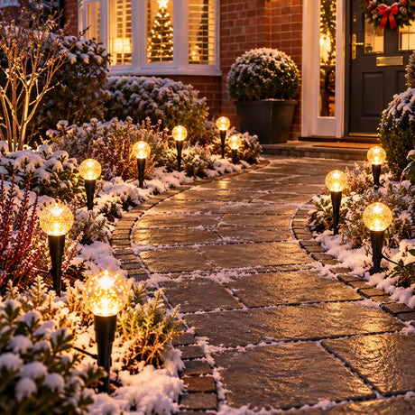 Outdoor Christmas string lights with glowing warm white globe bulbs mounted on black stakes, set in snow beside stone pavers in a front garden path.