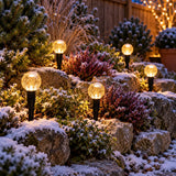 Outdoor garden pathway at dusk with several spherical solar path lights glowing warmly through snow-covered shrubs in a garden rockery