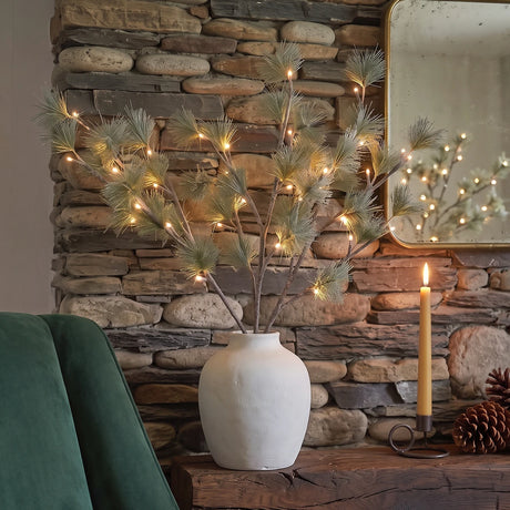 A decorative artificial pine branch arrangement with warm white string lights in a matte white ceramic vase, styled on a rustic wooden mantel in front of a stone fireplace with a lit candle and pinecones nearby.
