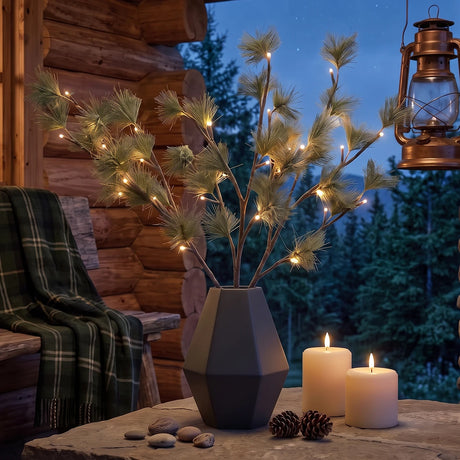 Faux pine branches with warm white string lights in a dark gray geometric vase, styled on a rustic table with candles, pinecones, and a cozy log cabin background at dusk.