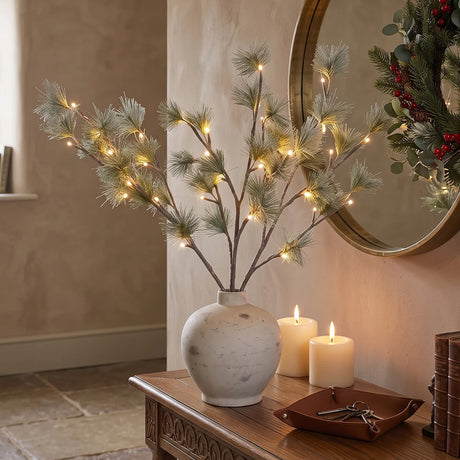 A decorative artificial pine branch arrangement with warm white string lights, displayed in a white ceramic vase