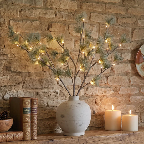 An artificial pine branch arrangement with warm white string lights in a round off-white ceramic vase, styled on a wooden shelf against a rustic stone wall with candles and books.