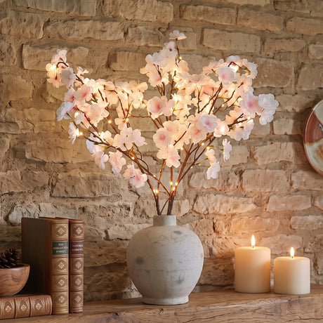 A vase of artificial cherry blossom branches with glowing warm white lights, displayed on a wooden shelf against a rustic stone wall, with ember candles and books nearby.