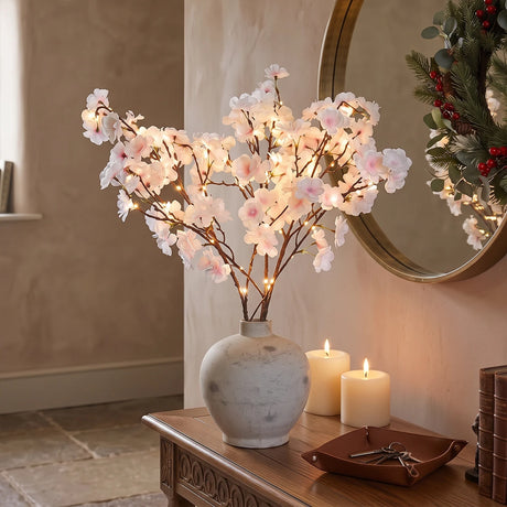 A decorative artificial cherry blossom branch arrangement with warm white string lights, displayed in a white ceramic vase on a wooden console table in a styled interior setting.
