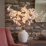 A decorative artificial cherry blossom branch arrangement with glowing warm lights, displayed in a matte white ceramic vase on a wooden mantel in front of a stone wall.