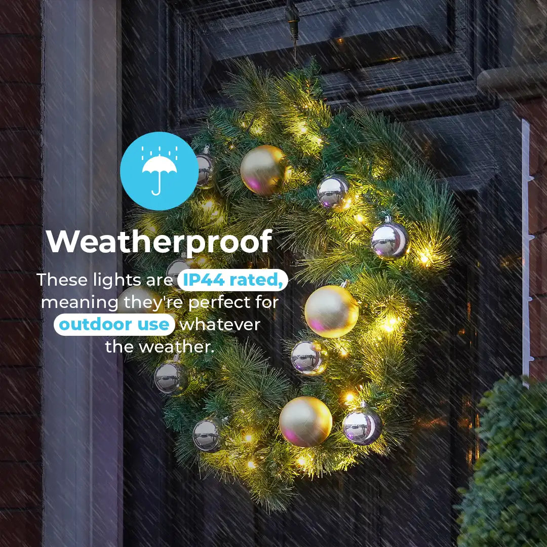 A green Christmas wreath with warm white lights and reflective gold and silver ornaments hanging on a dark front door in the rain. Text on the left says the lights are weatherproof IP44 rated for outdoor use.