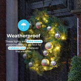 A green Christmas wreath with warm white lights and reflective gold and silver ornaments hanging on a dark front door in the rain. Text on the left says the lights are weatherproof IP44 rated for outdoor use.