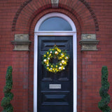 A green holiday wreath with warm white string lights and gold ornaments hanging on a black front door set within a red brick building.