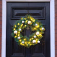 A green Christmas wreath with warm white lights, gold and silver ornaments, and a hanging loop, displayed on a black front door.