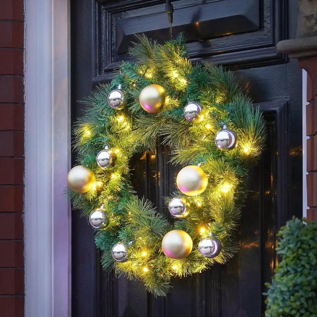 A green pine wreath decorated with warm white string lights and large metallic gold and silver Christmas ornaments, hanging on a dark front door.