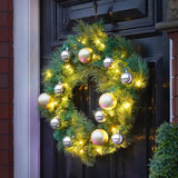 A green pine wreath decorated with warm white string lights and large metallic gold and silver Christmas ornaments, hanging on a dark front door.