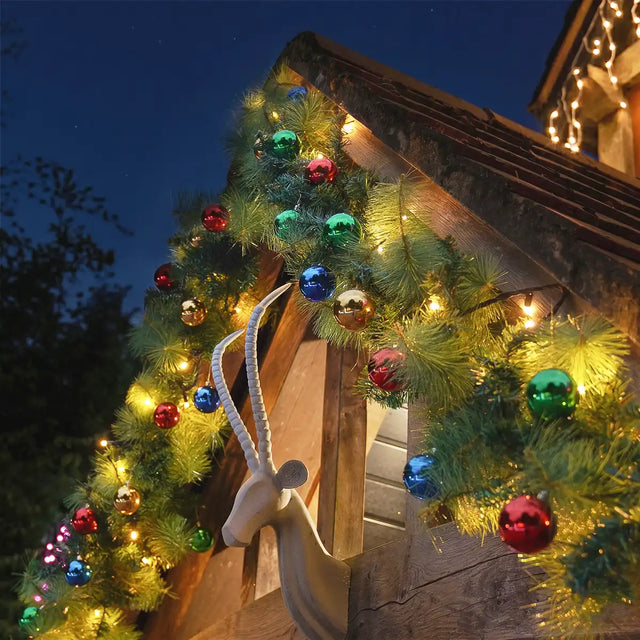 Outdoor Christmas garland wrapped around a wooden house corner at night, decorated with multicolored reflective ornaments, warm white string lights, and a white goat head wall decor.