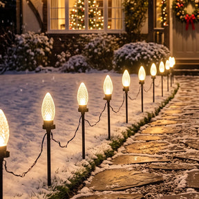 Outdoor Christmas landscape lights with glowing bulb-shaped bulbs mounted on black stakes along a snowy stone path in front of a house at night.