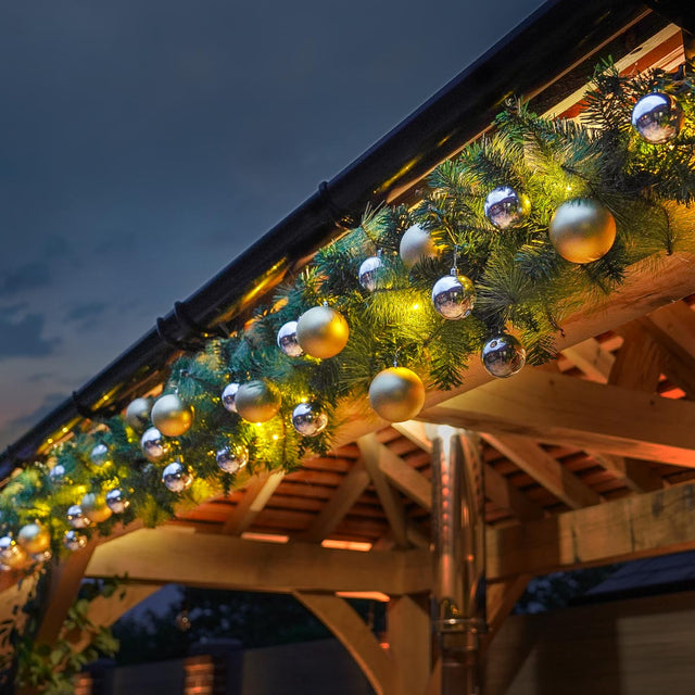Christmas garland draped over a wooden roof eave at dusk, decorated with green pine branches, warm white string lights, and reflective silver and gold ornaments.