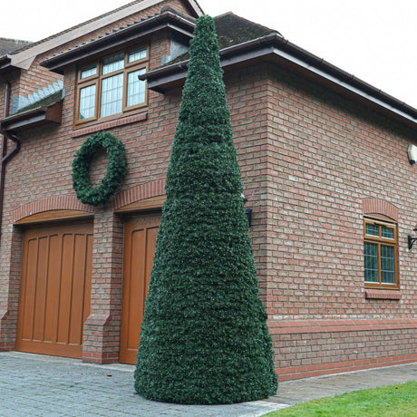 A tall, cone green topiary tree stands in front of a red brick house with wooden garage doors and a matching green wreath on the wall.