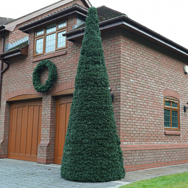 A tall, cone green topiary tree stands in front of a red brick house with wooden garage doors and a matching green wreath on the wall.