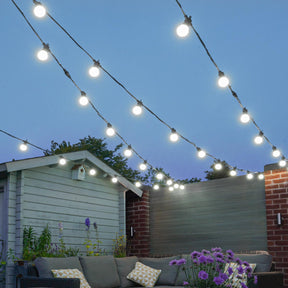 Outdoor festoon lights with glowing round bulbs suspended above a patio area, with a gray sofa, house, fence, and blue evening sky in the background.