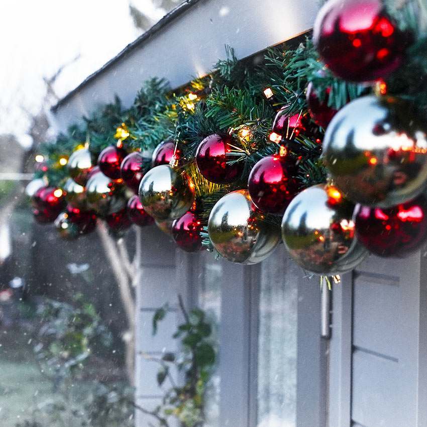 A festive holiday garland draped over a white exterior wall or door, decorated with green pine branches, warm white string lights, and shiny red and silver reflective ornaments.