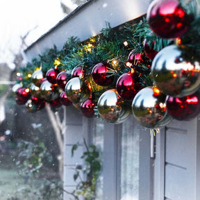 A festive holiday garland draped over a white exterior wall or door, decorated with green pine branches, warm white string lights, and shiny red and silver reflective ornaments.
