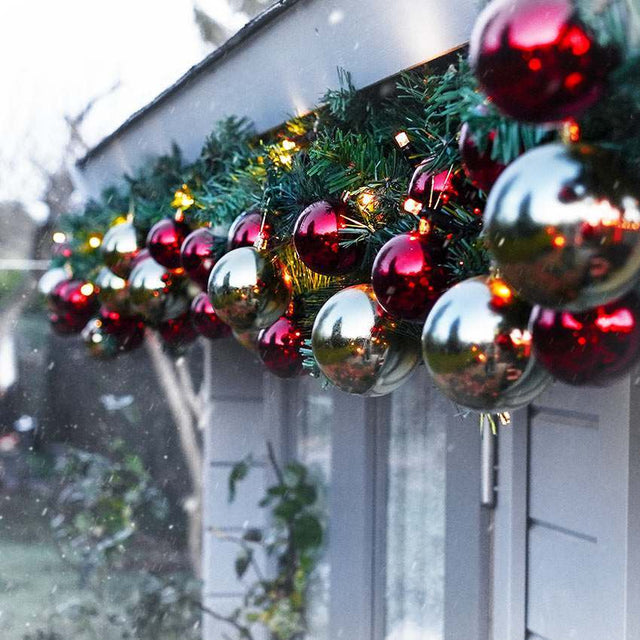 A festive holiday garland draped over a white exterior wall or door, decorated with green pine branches, warm white string lights, and shiny red and silver reflective ornaments.